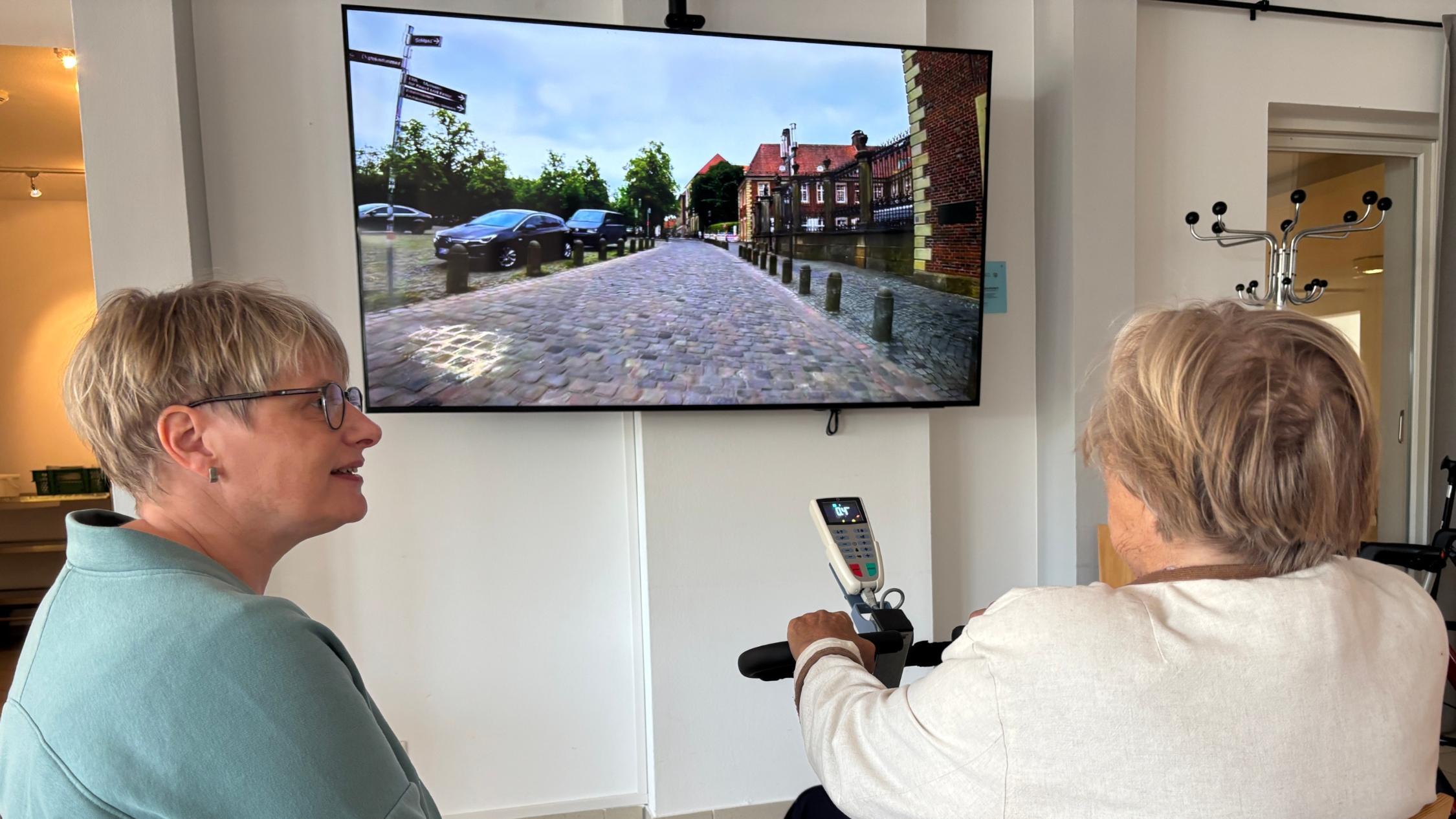 Eine Seniorin radelt auf einem Heimtrainer und verfolgt auf einem Bildschirm ihre virtuelle Radtour durch Münster, beobachtet von einer Mitarbeiterin des Sozialen Dienstes.